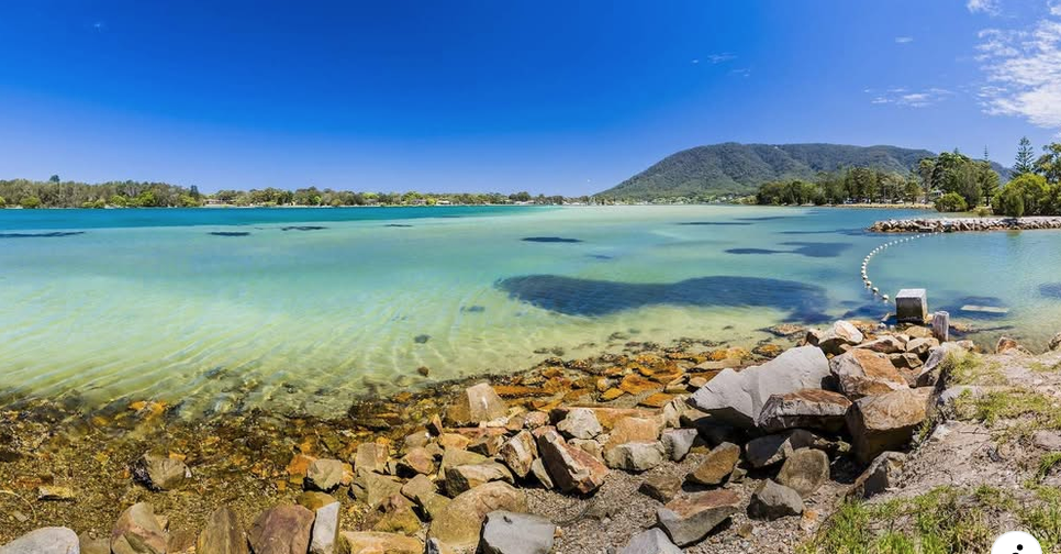 Panoramic view of a tranquil body of water with a rocky shoreline, surrounded by lush greenery and hills under a clear blue sky.